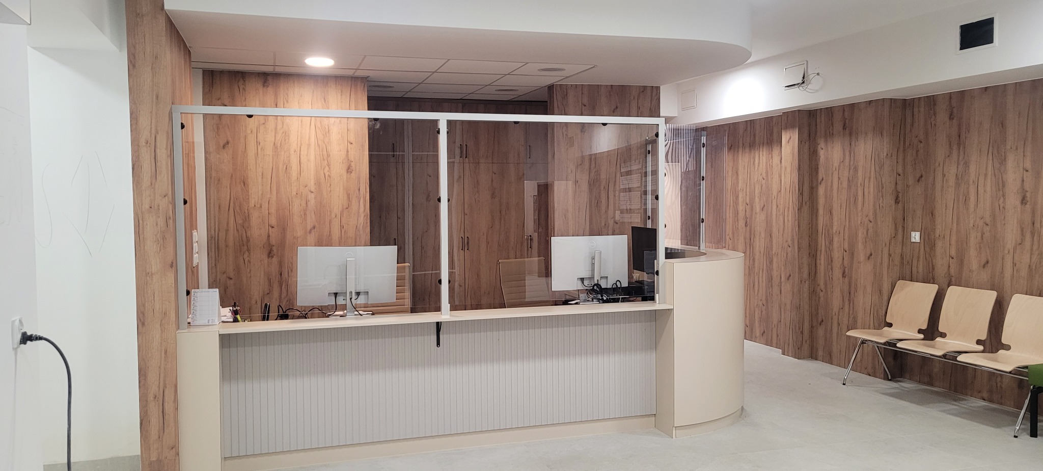 Nowoczesne wnętrze biurowe z drewnianymi panelami ściennymi, przeszklonym stanowiskiem recepcyjnym z dwoma monitorami i trzema krzesłami dla oczekujących gości.