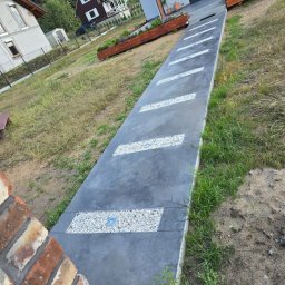 BETON TPS Sołtysiak Tomasz - Długa, szara ścieżka z wbudowanymi prostokątnymi wstawkami z białego żwiru i punktowym oświetleniem, prowadząca do garażu, otoczona trawnikiem i ozdobnymi skrzyniami z kwiatami.