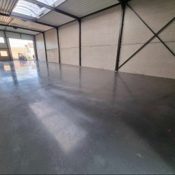 BETON TPS Sołtysiak Tomasz - Wnętrze hali przemysłowej z gładką, szarą posadzką przemysłową, widoczne stalowe elementy konstrukcyjne i wózek widłowy w oddali przez otwarte wrota.