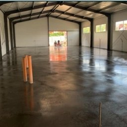 BETON TPS Sołtysiak Tomasz - Świeżo wylana, błyszcząca posadzka w hali przemysłowej z widocznymi rurami PCV przy ścianach i otwartą bramą w tle.