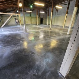 BETON TPS Sołtysiak Tomasz - Wnętrze hali z drewnianą konstrukcją dachu i świeżo wykonaną, ciemną posadzką. Na posadzce widoczne ślady po obróbce. W tle maszyna do polerowania posadzek.