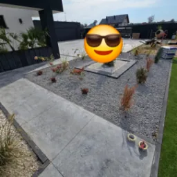 Nowoczesny taras z betonowych płyt w ogrodzie, obok rabata z szarym żwirem i roślinami, widoczne taśmy miernicze.