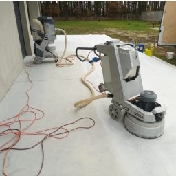 BETON TPS Sołtysiak Tomasz - Szlifowanie betonowej powierzchni za pomocą profesjonalnej szlifierki podłogowej z odkurzaczem przemysłowym, widoczne kable i węże, na zewnątrz budynku.