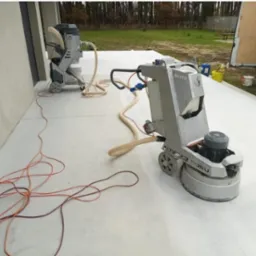 Szlifowanie betonowej powierzchni za pomocą profesjonalnej szlifierki podłogowej z odkurzaczem przemysłowym, widoczne kable i węże, na zewnątrz budynku.