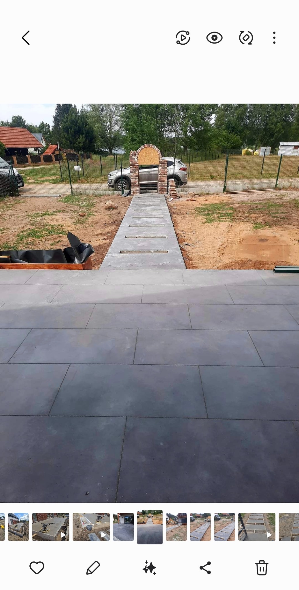 Nowoczesny taras z szarych płyt betonowych prowadzący do furtki z cegły, widok na ogród i zaparkowany samochód w tle.