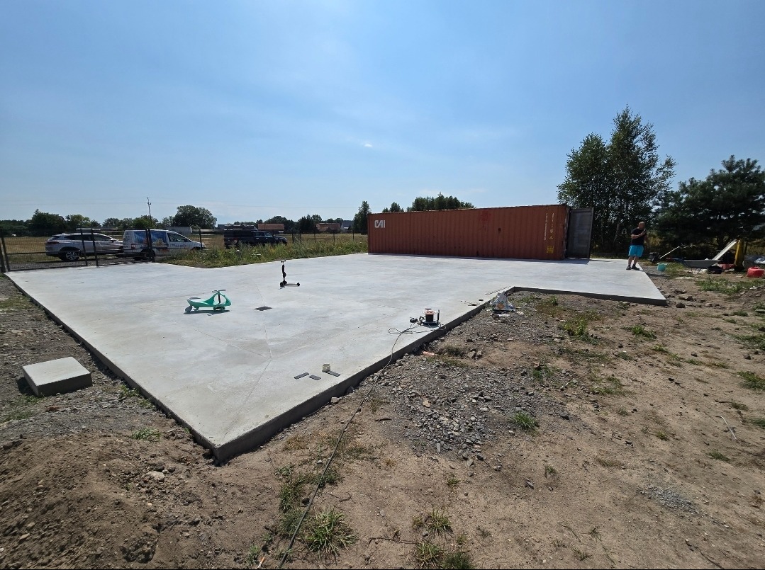 Świeżo wylana, szara płyta betonowa na zewnątrz, obok stoi kontener, widoczne zabawki dziecięce i samochody w tle.