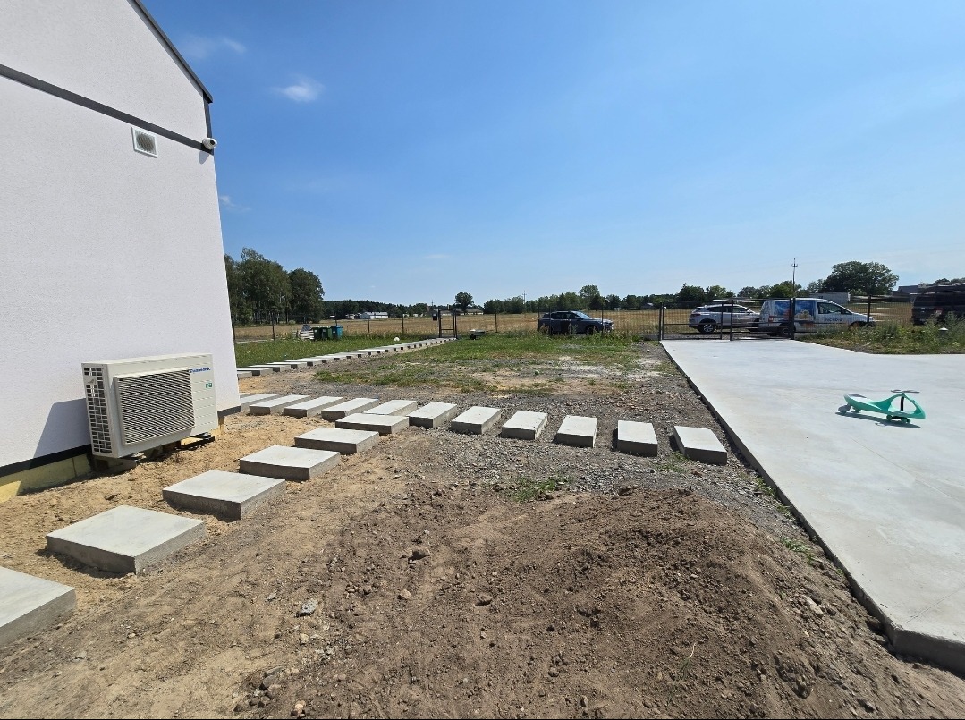 Ścieżka z betonowych płyt prowadząca od budynku do betonowego tarasu, w tle widoczne samochody i ogrodzenie.