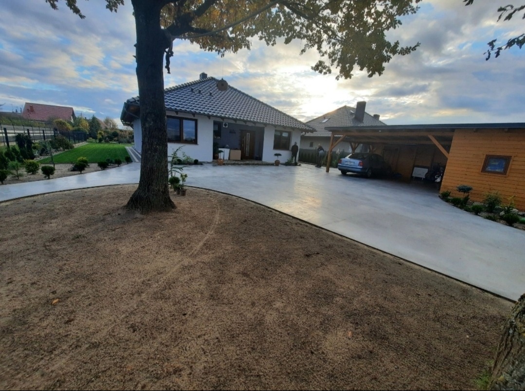 Nowa, gładka nawierzchnia betonowa podjazdu i ścieżki wokół domu jednorodzinnego z widocznym samochodem pod wiatą garażową, trawnikiem i drzewami w jesiennej scenerii.