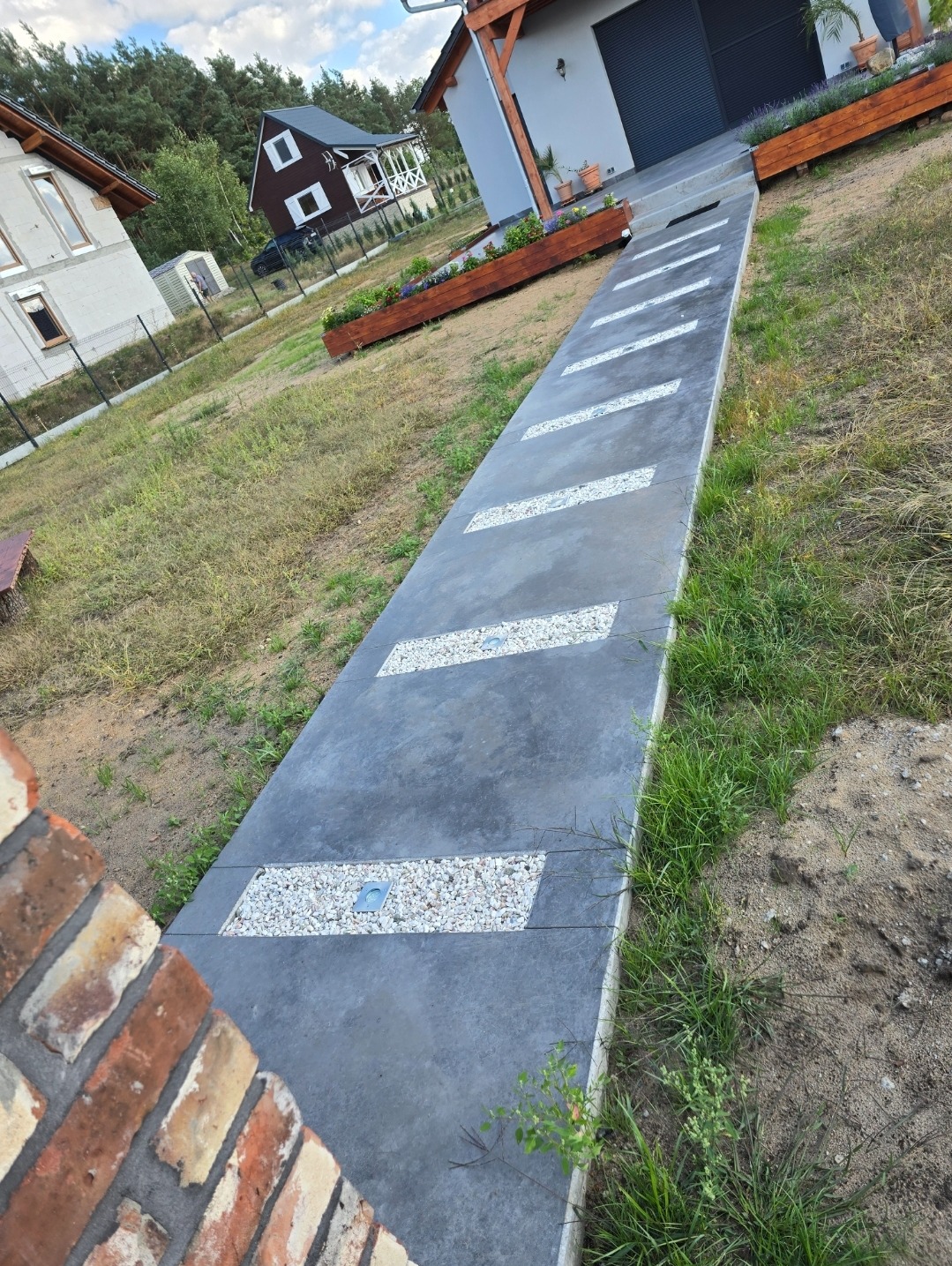 Długa, szara ścieżka z wbudowanymi prostokątnymi wstawkami z białego żwiru i punktowym oświetleniem, prowadząca do garażu, otoczona trawnikiem i ozdobnymi skrzyniami z kwiatami.