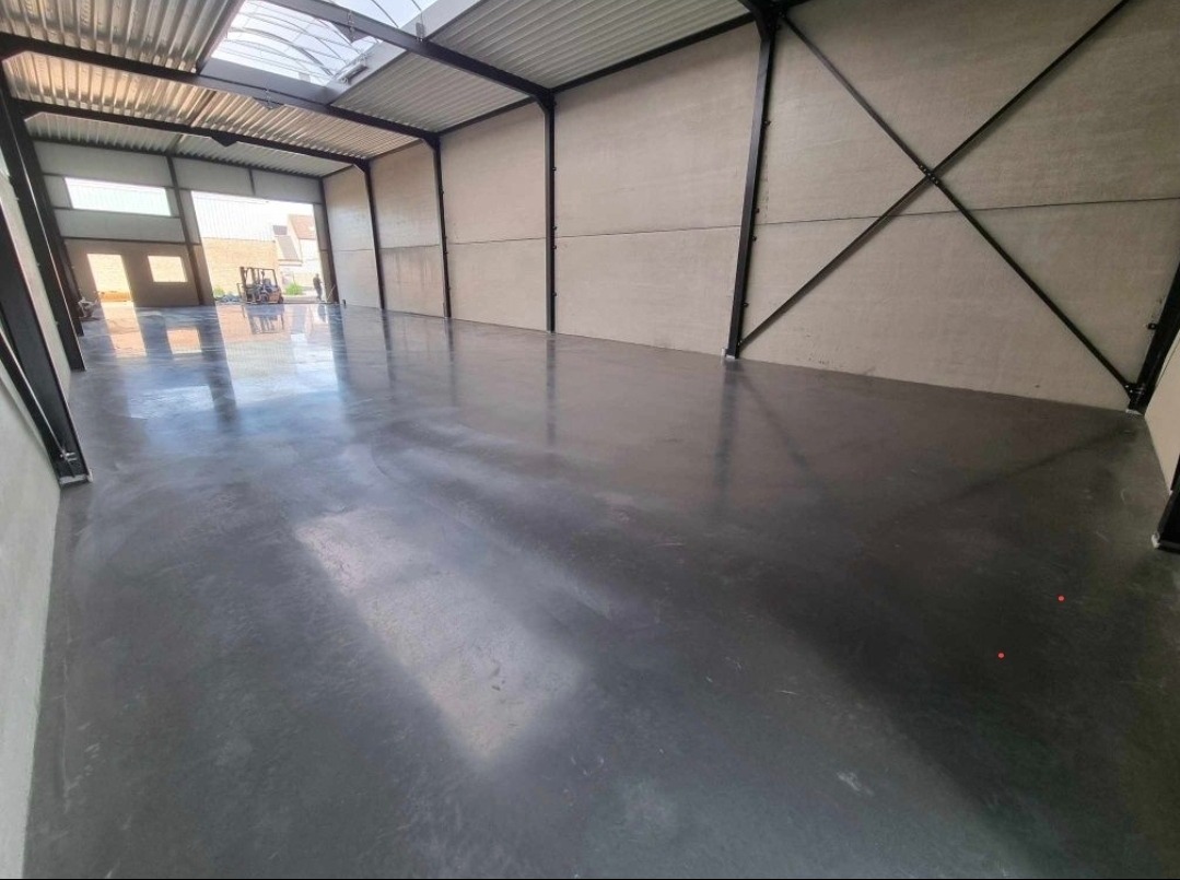 Wnętrze hali przemysłowej z gładką, szarą posadzką przemysłową, widoczne stalowe elementy konstrukcyjne i wózek widłowy w oddali przez otwarte wrota.