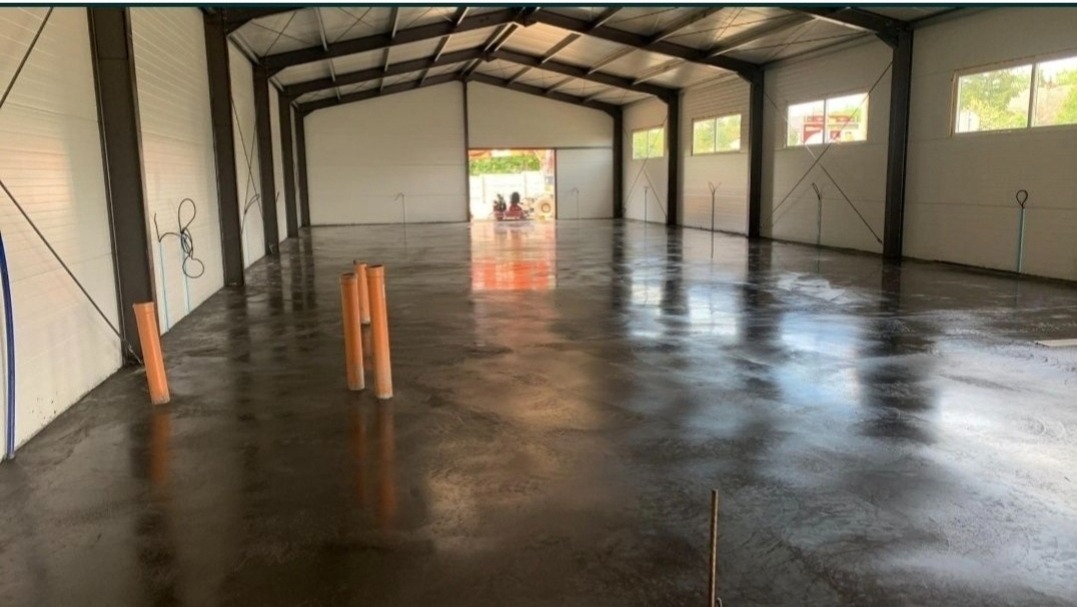 Świeżo wylana, błyszcząca posadzka w hali przemysłowej z widocznymi rurami PCV przy ścianach i otwartą bramą w tle.