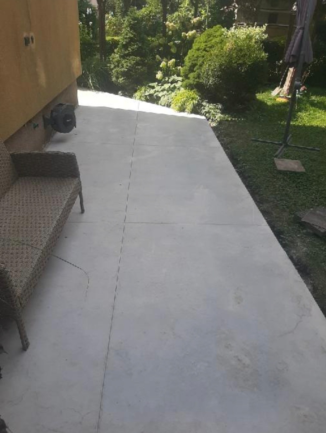 Świeżo wylany szary taras przylegający do budynku, z widocznym rattanowym siedziskiem i zielenią w tle.