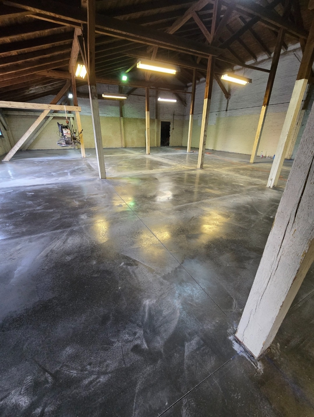 Wnętrze hali z drewnianą konstrukcją dachu i świeżo wykonaną, ciemną posadzką. Na posadzce widoczne ślady po obróbce. W tle maszyna do polerowania posadzek.