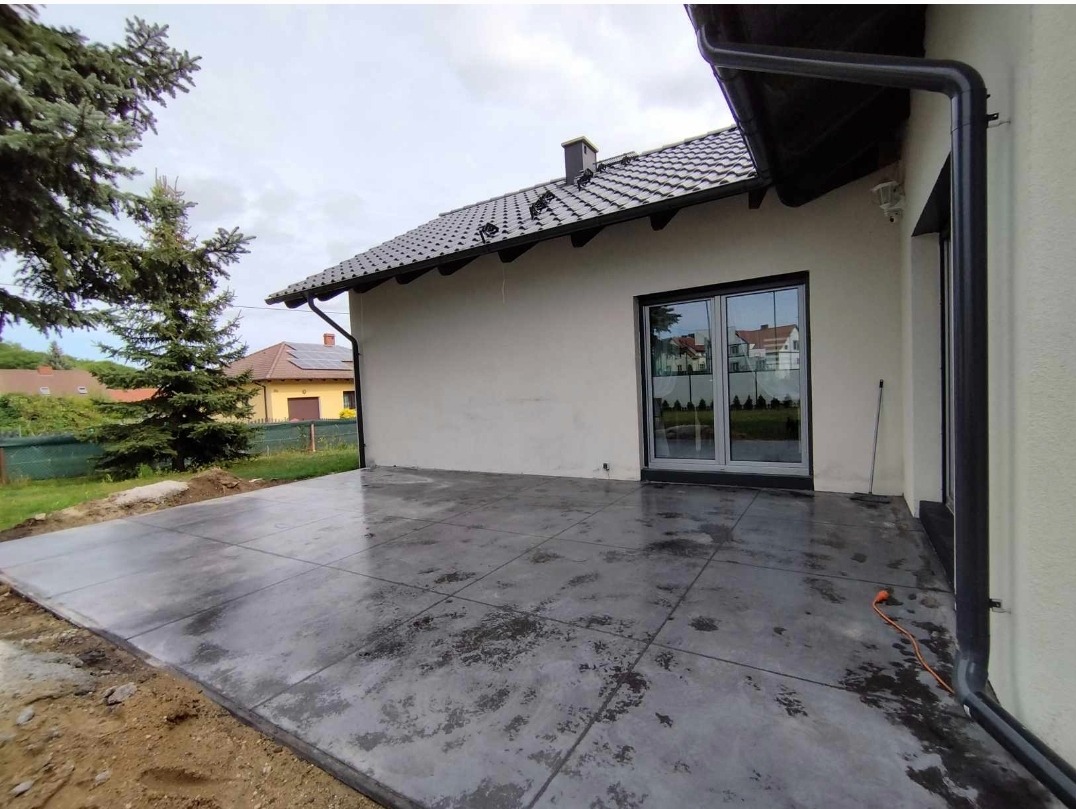 Wykończony taras z betonu architektonicznego w Bobrowicach, z widocznymi śladami wilgoci i delikatną teksturą, przylegający do budynku z przesuwnymi drzwiami balkonowymi i czarną rynną.