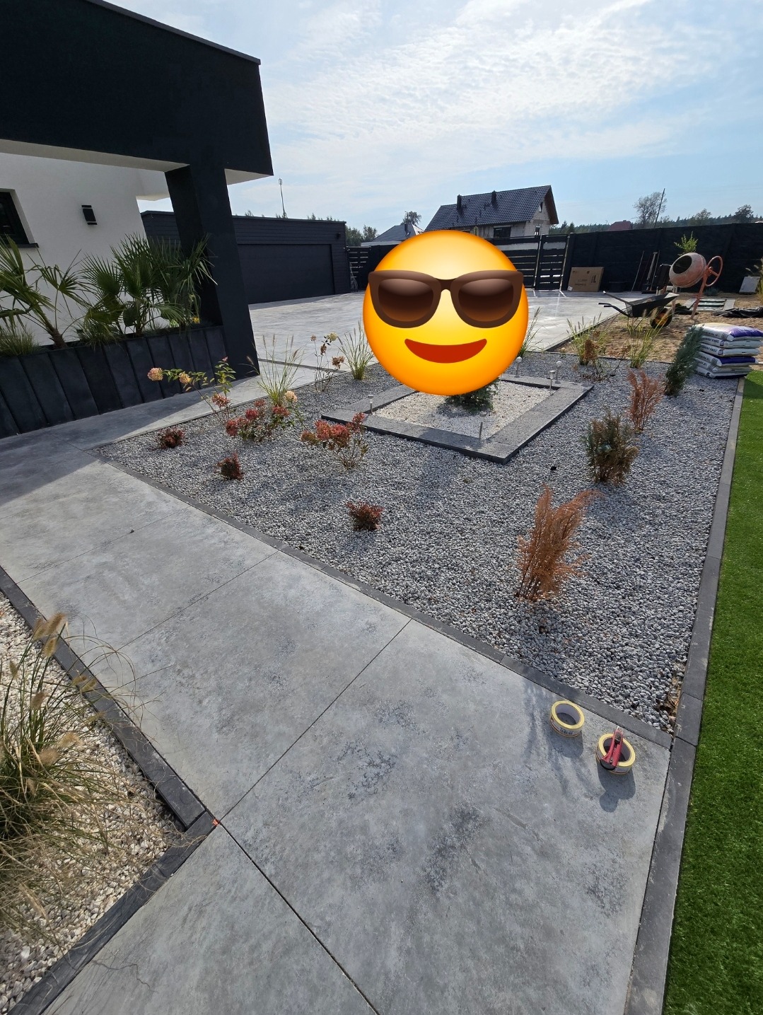 Nowoczesny taras z betonowych płyt w ogrodzie, obok rabata z szarym żwirem i roślinami, widoczne taśmy miernicze.