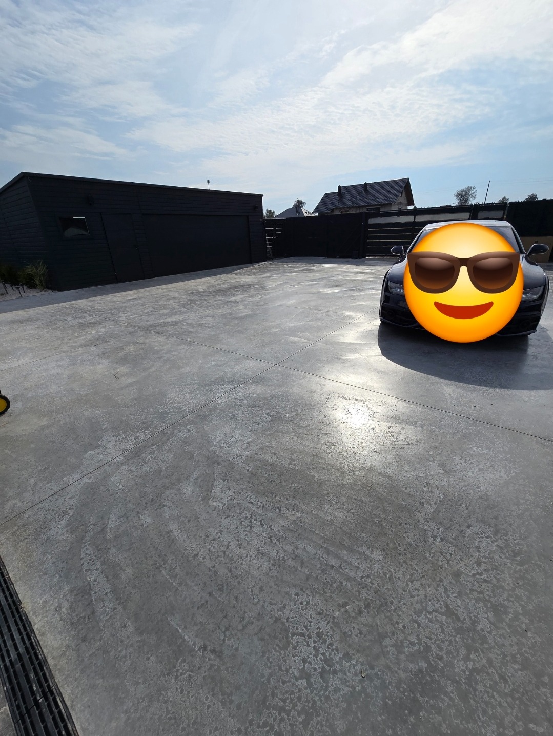 Szara, gładka nawierzchnia przed garażem i domem w słoneczny dzień, z widocznym samochodem z naklejoną emotikoną na przedniej szybie.