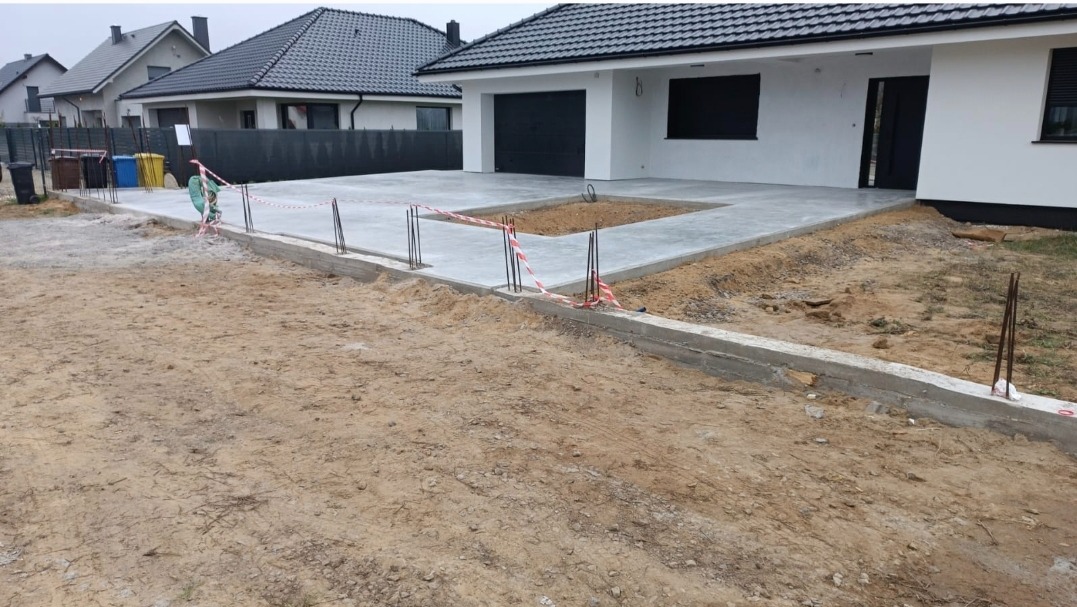 Wylewka betonowa tarasu przed domem w trakcie realizacji, widoczne pręty zbrojeniowe i taśmy ostrzegawcze, w tle dom z czarną stolarką okienną i bramą garażową.