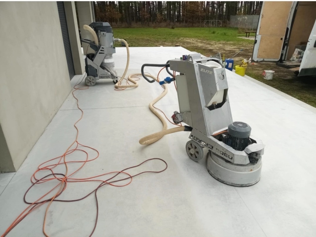 Szlifowanie betonowej powierzchni za pomocą profesjonalnej szlifierki podłogowej z odkurzaczem przemysłowym, widoczne kable i węże, na zewnątrz budynku.