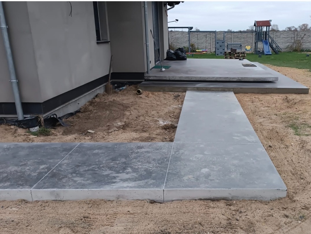 Nowoczesny taras z szarego betonu architektonicznego w trakcie budowy, widoczny fragment ścieżki z płyt betonowych prowadzącej do podwyższonej części tarasu przy domu jednorodzinnym, otoczenie...