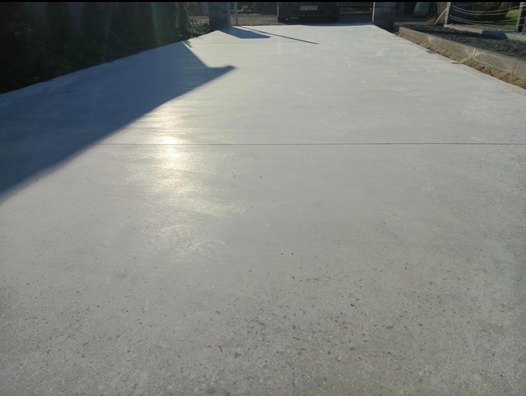 Świeżo wylana, gładka powierzchnia z betonu, lekko połyskująca w słońcu, z widocznym cieniem drzewa po lewej stronie i samochodem w tle.