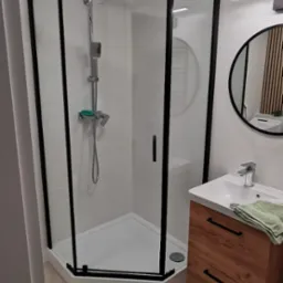 Nowoczesna łazienka z kabiną prysznicową narożną z czarnymi ramami, umywalką z szafką z drewnopodobnym frontem i okrągłym lustrem z drewnianymi lamelami w tle.