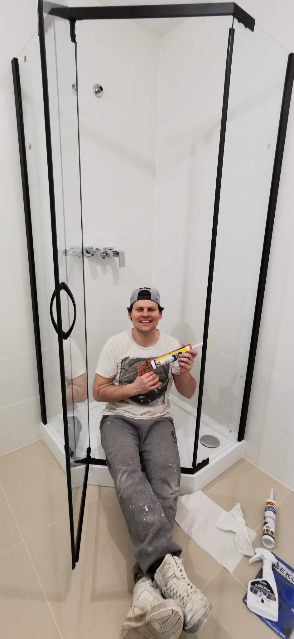 Uśmiechnięty mężczyzna w czapce i ubrudzonych farbą spodniach siedzi w narożnej kabinie prysznicowej z czarnymi ramami, trzymając tubę silikonu i pistolet do silikonu. Obok leżą narzędzia...