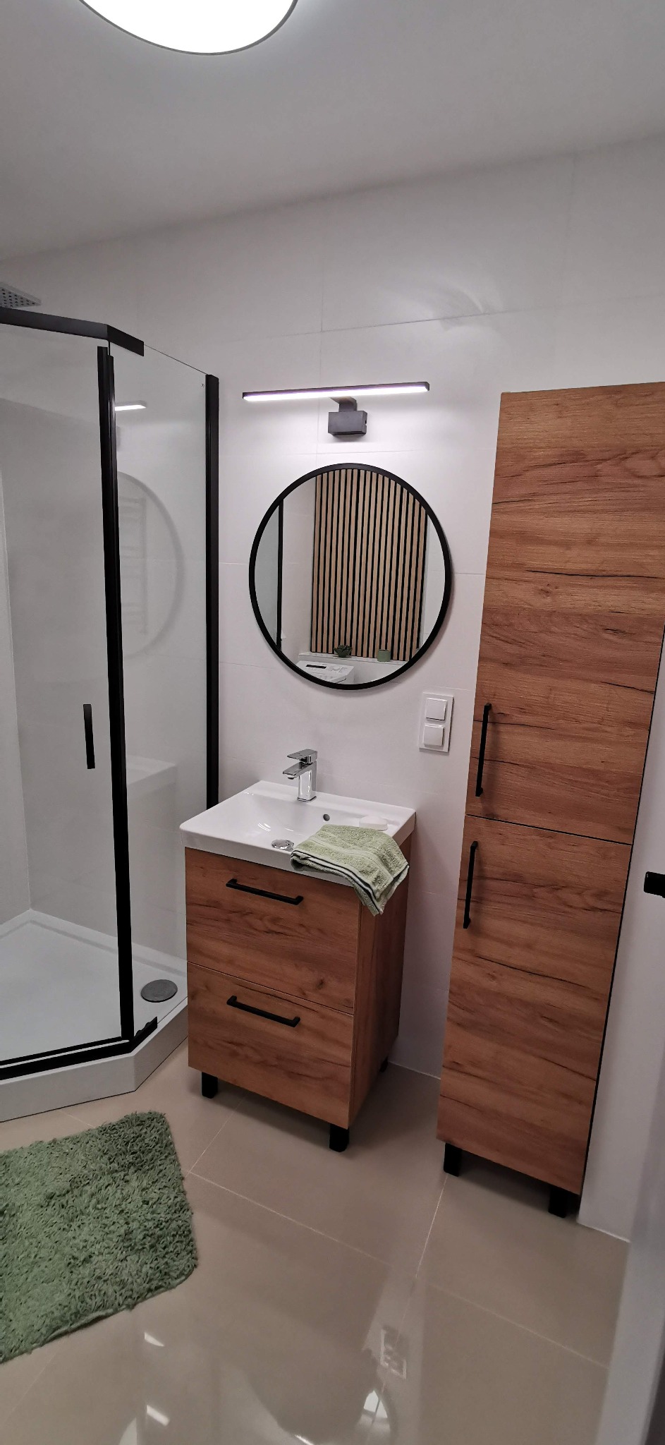 Nowoczesna łazienka z kabiną prysznicową typu walk-in, szafką z umywalką o drewnianym wykończeniu, okrągłym lustrem z podświetleniem LED oraz wysoką szafką, dopełniona zielonym dywanikiem.