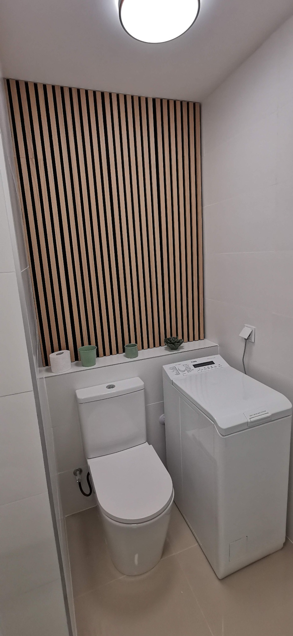 Nowoczesna łazienka z toaletą, pralką ładowaną od góry i dekoracyjną ścianą z drewnianych listew, dopełniona minimalistycznymi akcentami w postaci doniczek i rolki papieru toaletowego.
