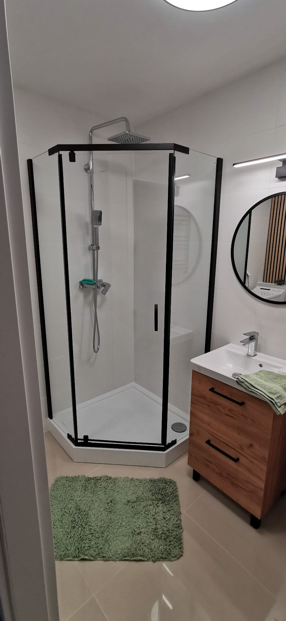 Nowoczesna łazienka z kabiną prysznicową narożną z czarnymi ramami, umywalką z szafką z drewnopodobnym frontem i okrągłym lustrem z drewnianymi lamelami w tle.