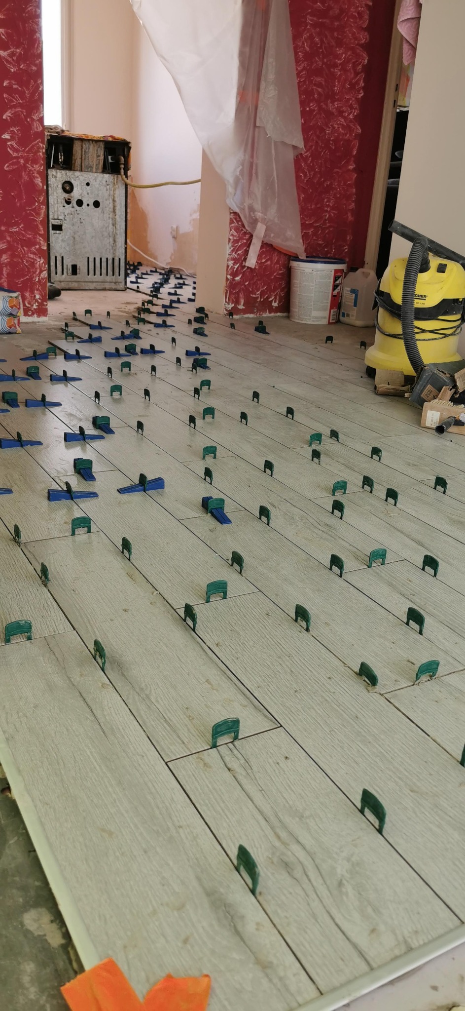 Ułożone panele podłogowe z jasnego drewna zabezpieczone klipsami poziomującymi w trakcie prac wykończeniowych; w tle widoczny odkurzacz przemysłowy i resztki materiałów budowlanych.
