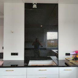 Lux-Kam Kamieniarstwo Łukasz Szewczyk - Czarny, błyszczący blat kamienny z wbudowanym zlewozmywakiem i panelem ściennym z tego samego materiału, widoczne złote uchwyty szafek i odbicie fotografa w panelu ściennym.