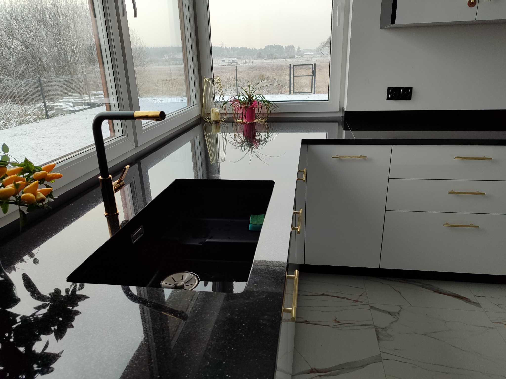 Czarny, błyszczący blat kuchenny z czarnym zlewem i złotą baterią na tle okna z zimowym krajobrazem, uzupełniony białymi szafkami z złotymi uchwytami i marmurową podłogą.