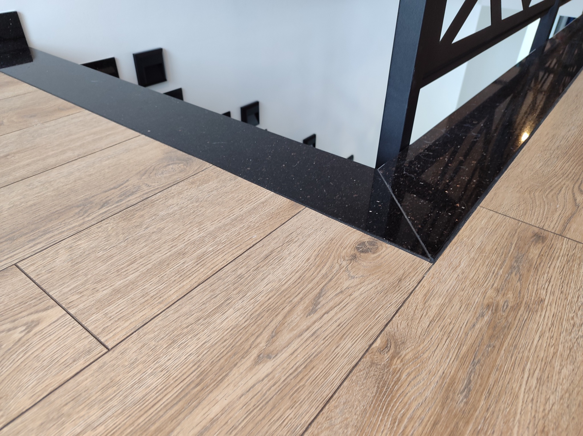 Fragment balustrady schodowej z czarnego kamienia, połączonej z drewnopodobną podłogą, widok z góry na schody w tle.
