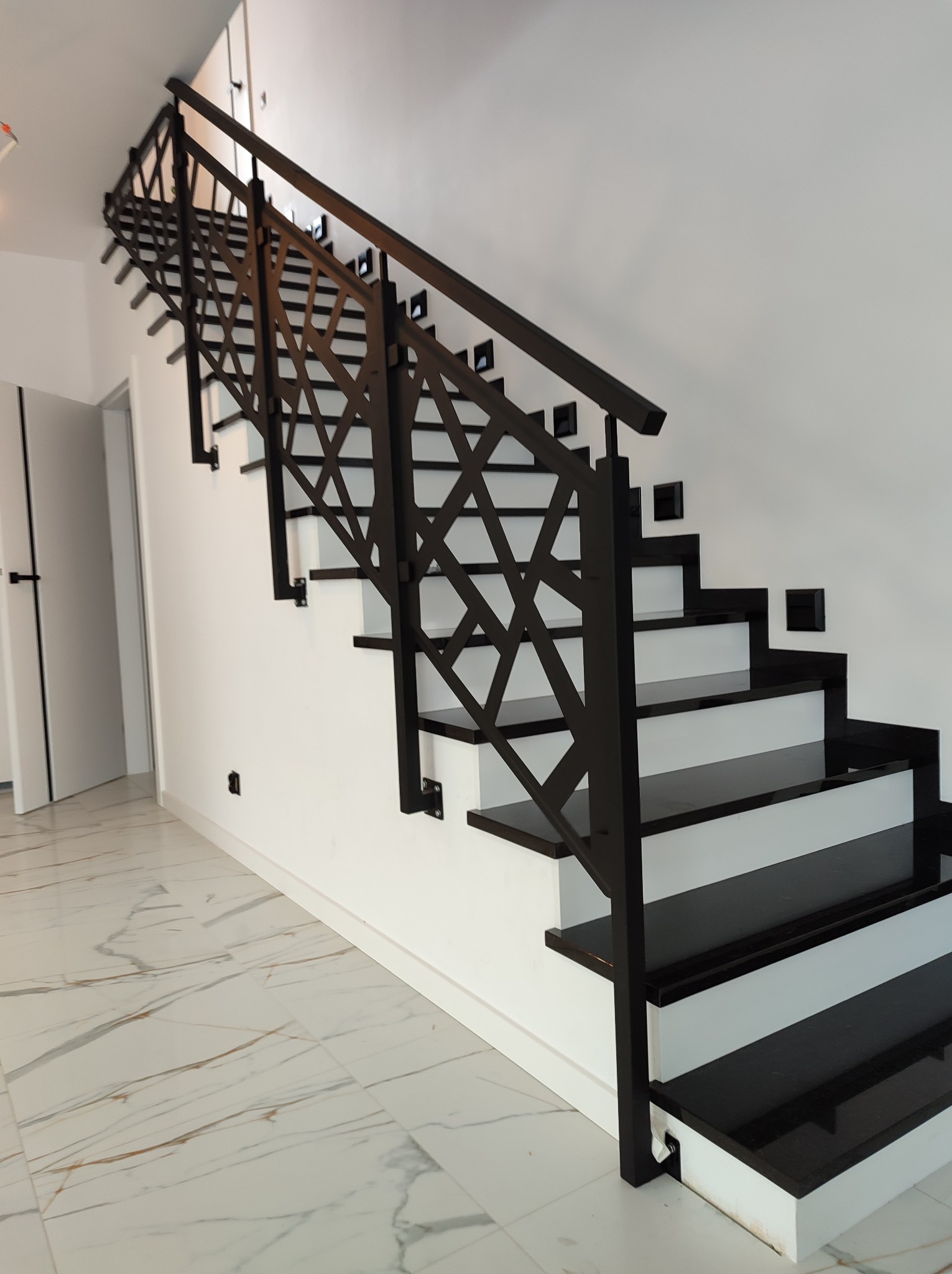 Nowoczesne schody wewnętrzne z czarnymi stopniami i geometryczną, czarną balustradą na tle białych ścian i marmurowej podłogi.