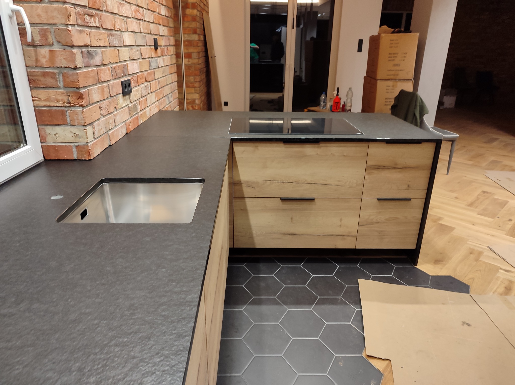 Szary blat kuchenny z wbudowanym zlewem i płytą indukcyjną, drewniane szafki, heksagonalne płytki podłogowe, fragment ceglanej ściany w tle.