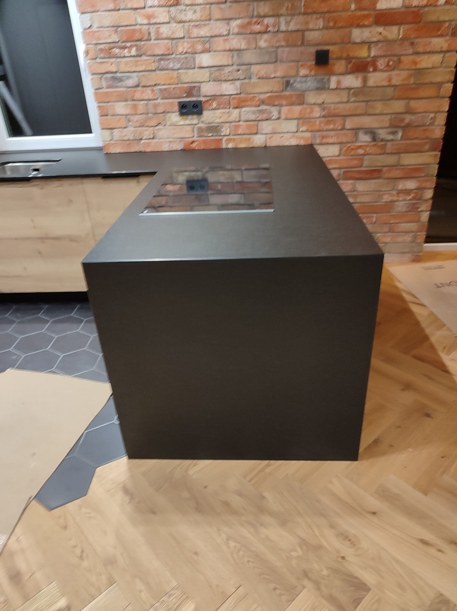 Ciemny, matowy blok kuchenny z wbudowaną płytą grzewczą, na tle ceglanej ściany i drewnianej podłogi w jodełkę, z widocznymi fragmentami kartonowych zabezpieczeń na podłodze.