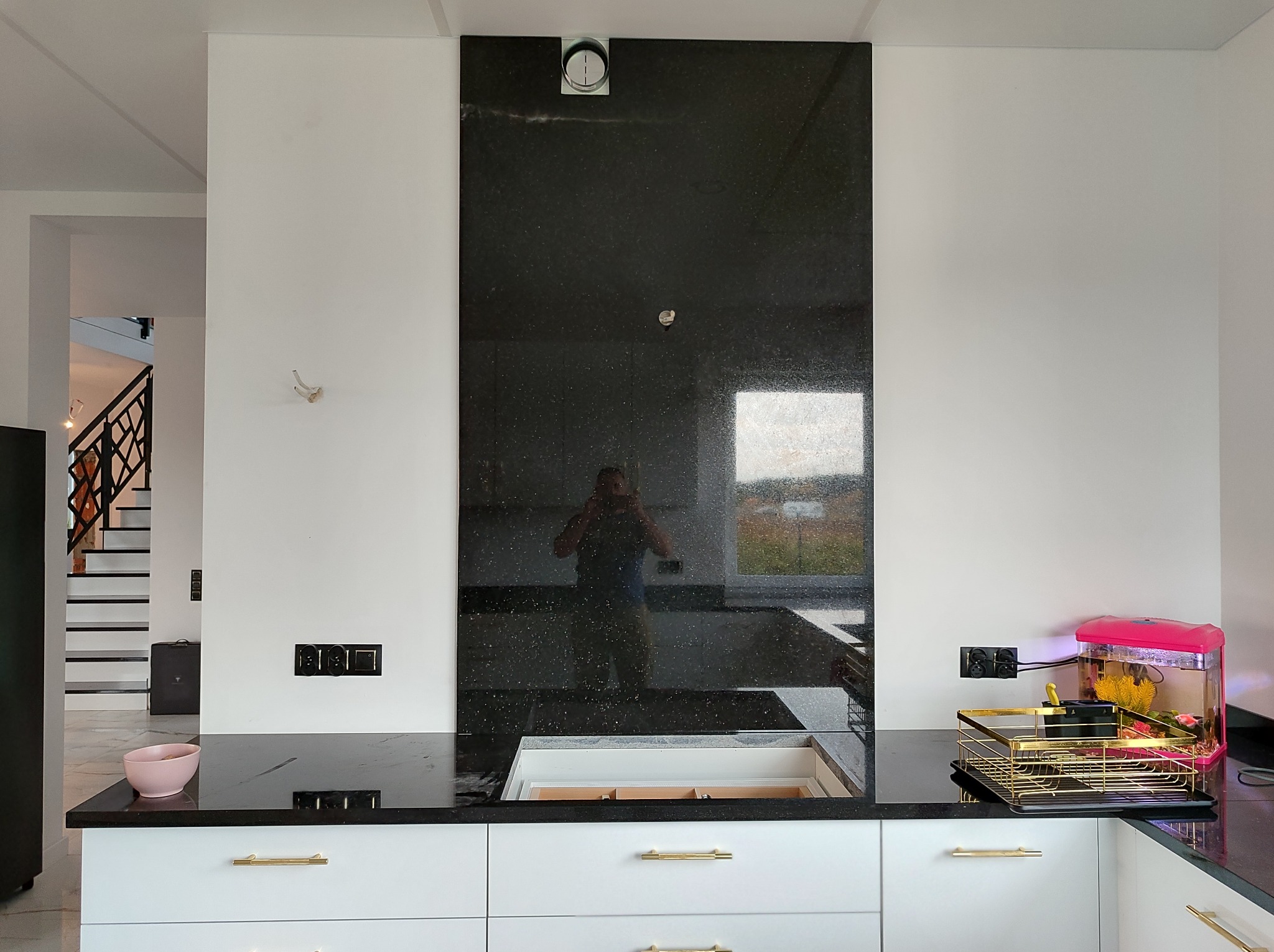 Czarny, błyszczący blat kamienny z wbudowanym zlewozmywakiem i panelem ściennym z tego samego materiału, widoczne złote uchwyty szafek i odbicie fotografa w panelu ściennym.