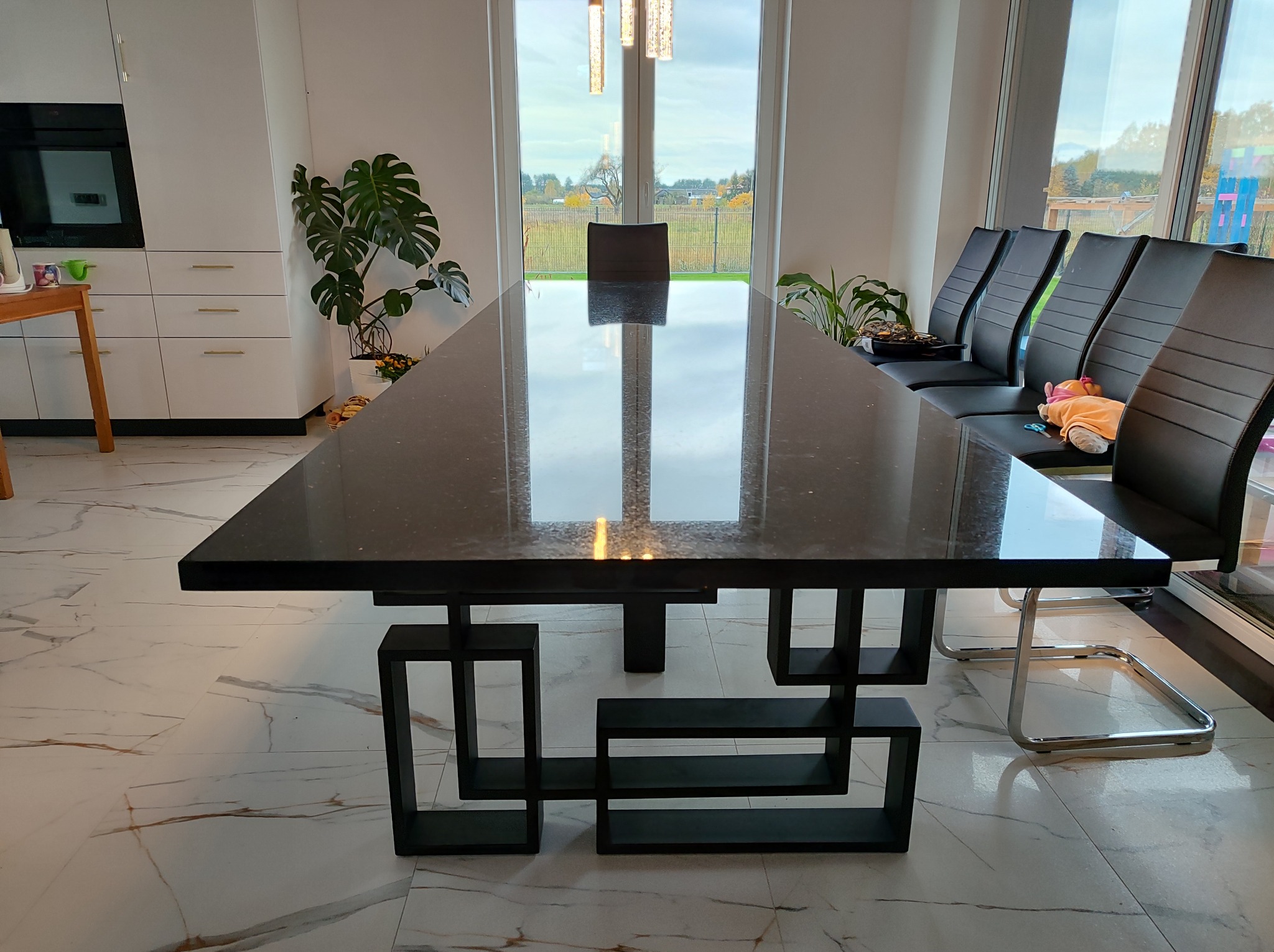 Długi, czarny stół jadalny z blatem o marmurkowej fakturze, wsparty na geometrycznej podstawie z czarnych, kwadratowych profili. W tle widoczne krzesła z czarnej skóry i duże okno z widokiem...