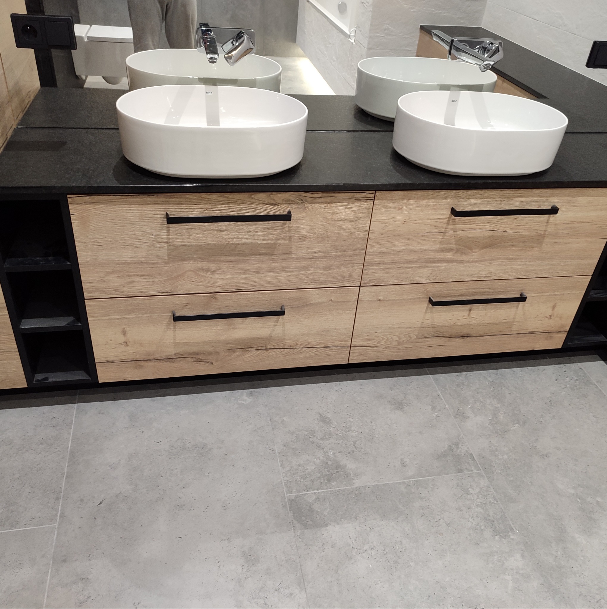 Dwie białe, okrągłe umywalki nablatowe na czarnym blacie z szafką z drewnopodobnymi frontami i czarnymi uchwytami, widok na szarą podłogę z płytek.