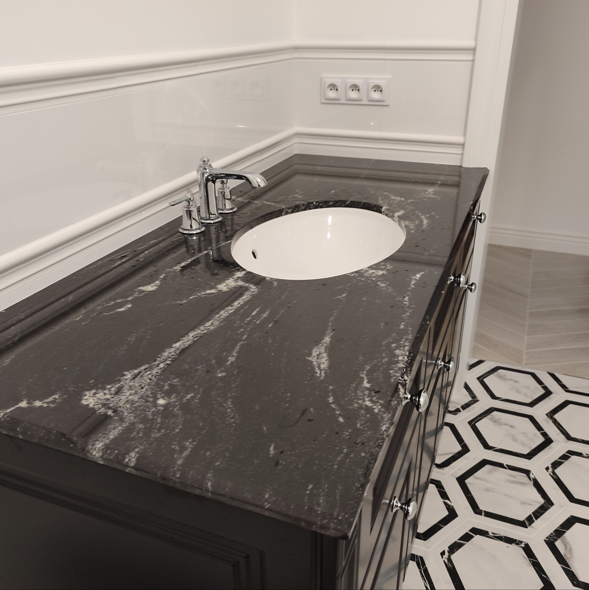 Czarny blat z białymi żyłkami, umywalka wpuszczana, chromowana bateria trzyotworowa, dekoracyjna listwa ścienna i heksagonalna podłoga w łazience.