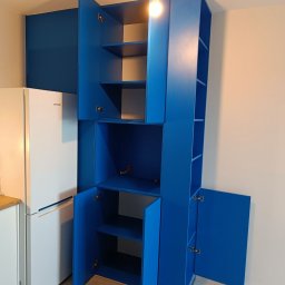 HiWall - Meble na Wymiar Wrocław - Wysoka, niebieska zabudowa meblowa z otwartymi szafkami w kuchni, obok biała lodówka Samsung, widoczna żarówka na suficie.