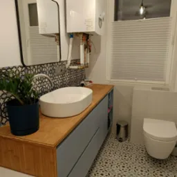 Nowoczesna łazienka z drewnianym blatem, umywalką nablatową, wzorzystymi płytkami i wiszącym piecem gazowym nad umywalką.