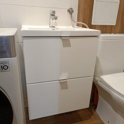 HiWall - Meble na Wymiar Wrocław - Biała umywalka z szafką podwieszaną w łazience, obok pralka i toaleta, widoczne drewnopodobne panele na ścianie.