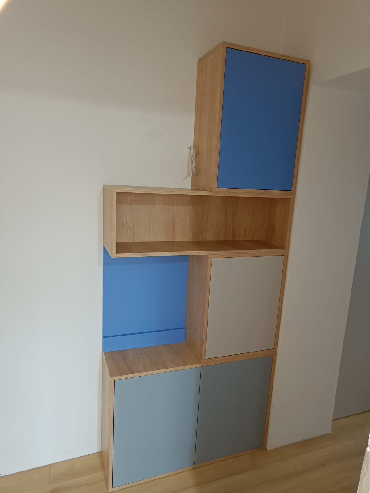 Wielopoziomowa zabudowa meblowa z elementami w kolorach: drewnopodobnym, niebieskim i szarym, dopasowana do skosu ściany.