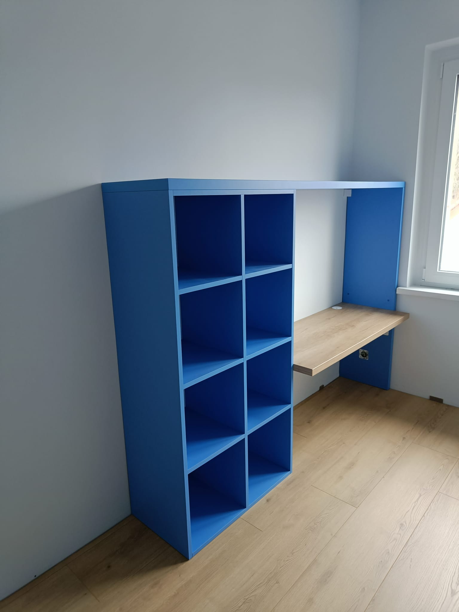 Niebieska zabudowa meblowa z regałem i biurkiem z drewnianym blatem, przymocowana do ściany w pokoju, widoczna z boku.