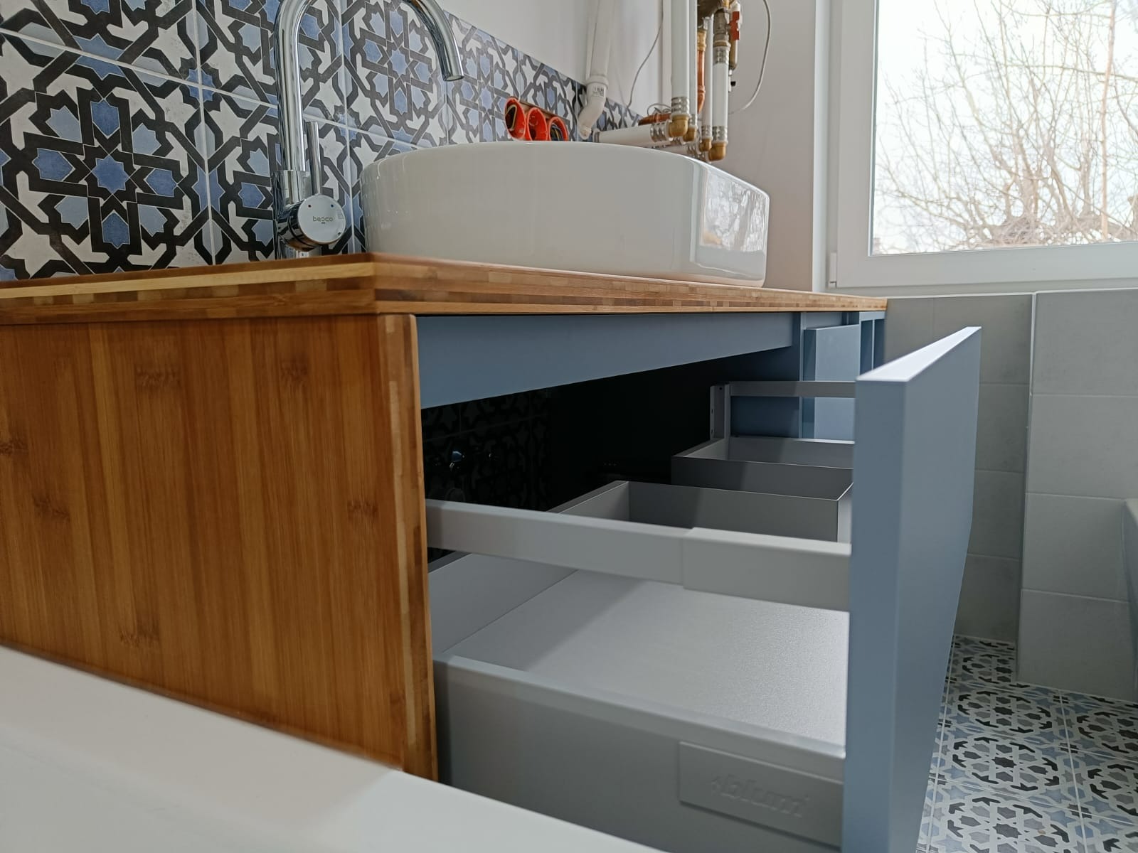 Łazienkowa szafka pod umywalkę z blatem z drewna bambusowego i szufladami w kolorze gołębim, otwarte szuflady z systemem Blum, mozaika na ścianie i podłodze.