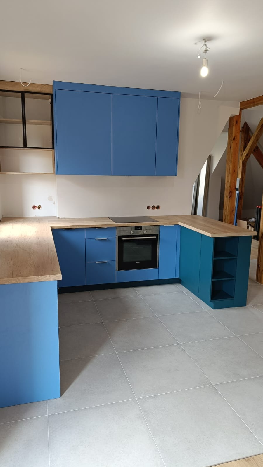 Niebieska zabudowa kuchenna z drewnianym blatem i piekarnikiem, widok na fragment poddasza z drewnianą konstrukcją dachową, szare płytki na podłodze.