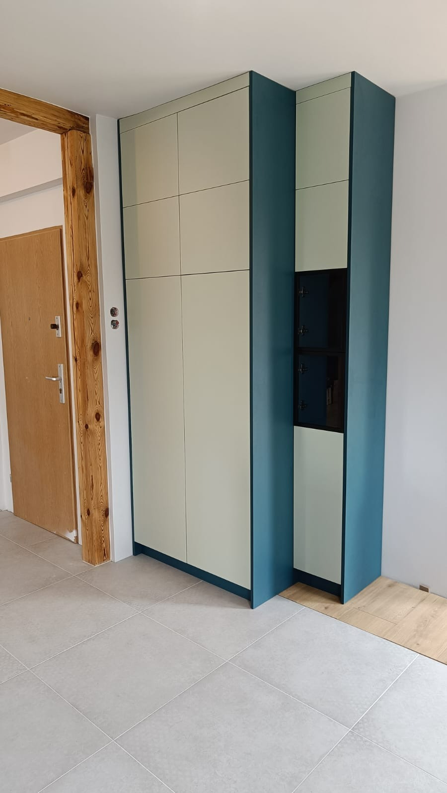 Szafa na wymiar z frontami w kolorze kremowym i obudową w odcieniu turkusowym, z wbudowanymi czarnymi szafkami, na tle jasnych płytek podłogowych i drewnianej futryny drzwi.