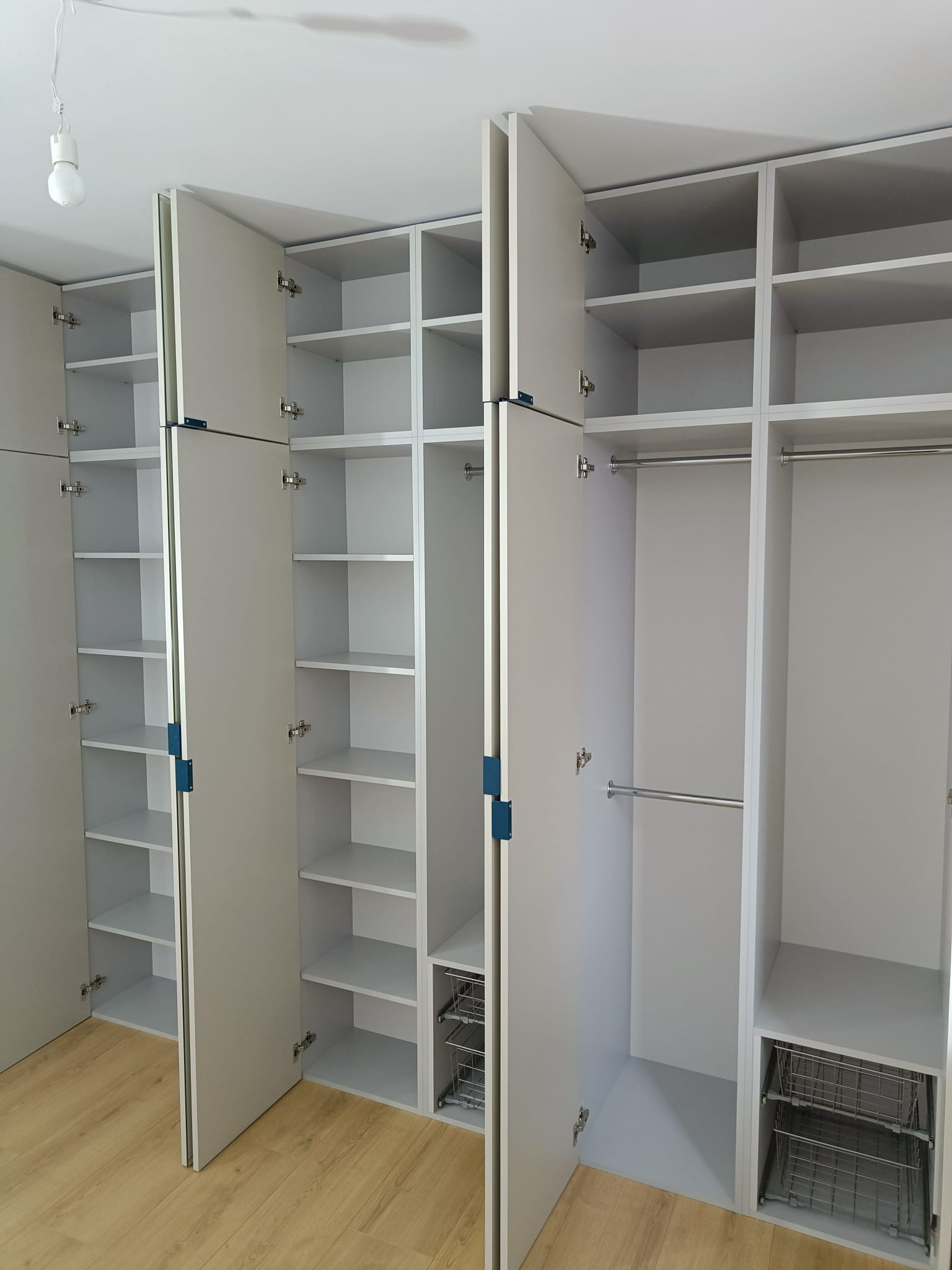 Jasnoszara garderoba z otwartymi drzwiami, widoczne półki, drążki na ubrania i wysuwane kosze druciane, na podłodze panele.
