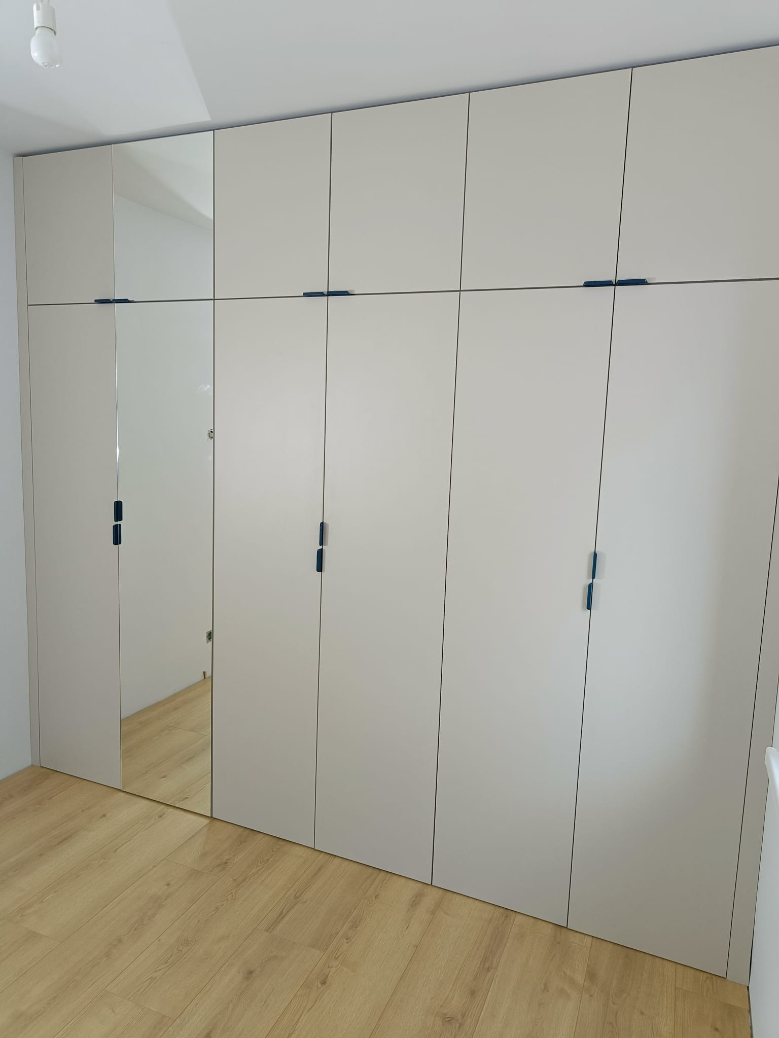 Biała, zabudowana szafa z frontami na wysoki połysk i ciemnymi, minimalistycznymi uchwytami, jeden segment z lustrzanymi drzwiami.